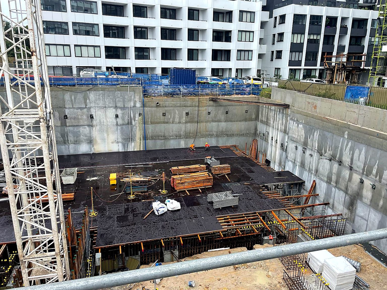 Workers installing black deck formwork at the base of a deep concrete-walled excavation.