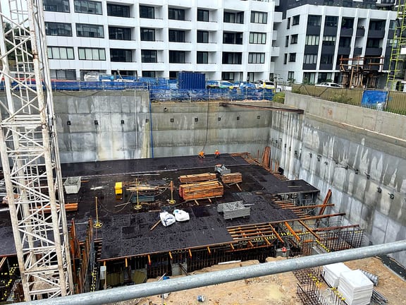 Workers installing black deck formwork at the base of a deep concrete-walled excavation.