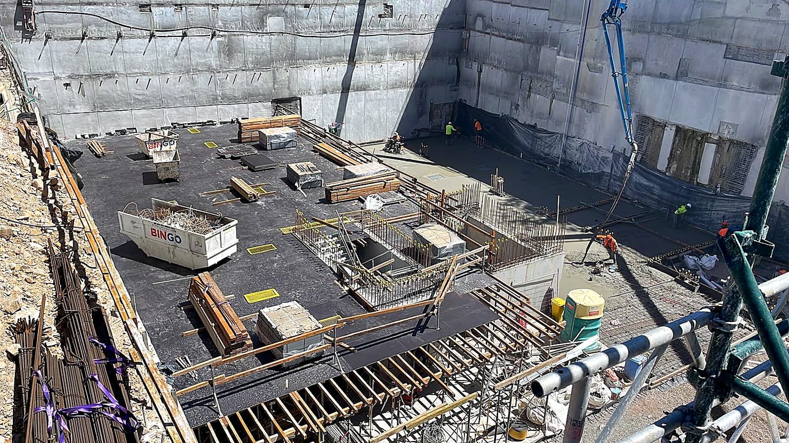 Workers placing and finishing concrete beside deck formwork in a deep basement excavation.