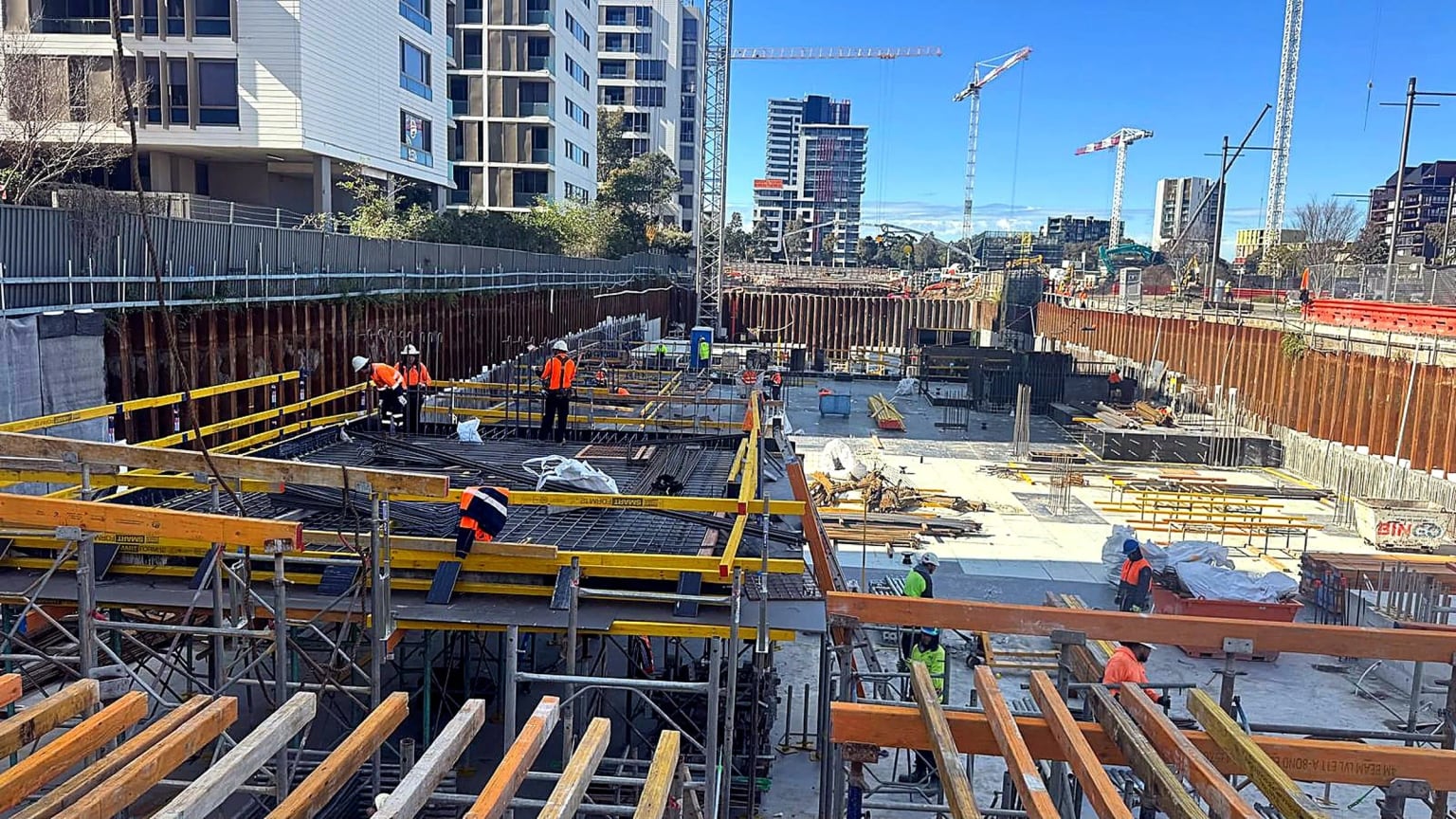 Site crew installing reinforcement and slab formwork in a deep urban excavation surrounded by apartment buildings.
